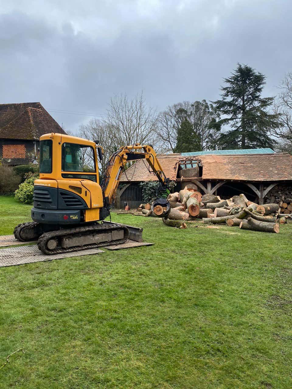 This is a photo of a tree which has grown through the roof of a barn that is being cut down and removed. There is a digger that is removing sections of the tree as well. Keyworth Tree Surgeons